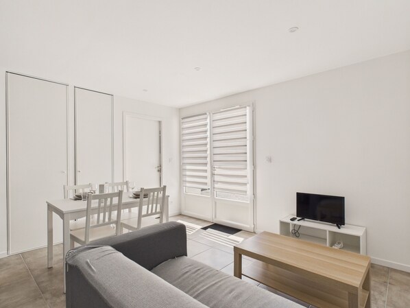 A minimalist living room with a sofa, a dining table with chairs, a TV on a stand, and a sliding door with blinds. - Apartment for 2 in St-Jean-de-Mont with garden (Saint-Jean-de-Monts)