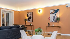 A modern living room with a glass coffee table, white chairs, a dark sofa, and two paintings on the walls.