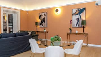 A modern living room with a glass coffee table, white chairs, a dark sofa, and two paintings on the walls.