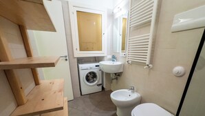 A compact bathroom with a washing machine, sink, bidet, and wooden shelves.