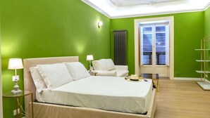 A room with a large bed, two bedside tables, a chair, a radiator, a window with blinds, and a shelf.