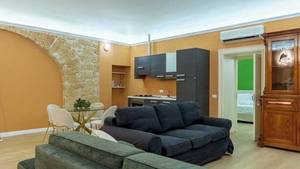 A living room with a dark sofa, a dining table with chairs, a kitchen area with cabinets, and a stone accent wall.