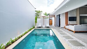 A modern indoor pool area with a clear pool, white lounge chairs, and a seating area with a sofa and TV. - The Moringa Bukit Villas (Kutuh)