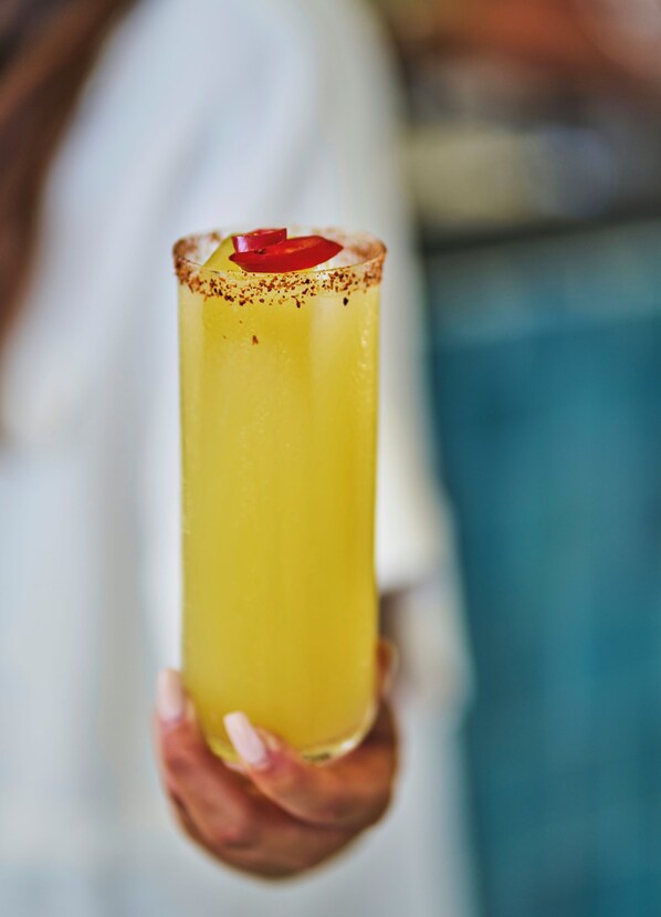 A person holding a tall glass with a yellow beverage, garnished with a red fruit and a sprinkle of spices.