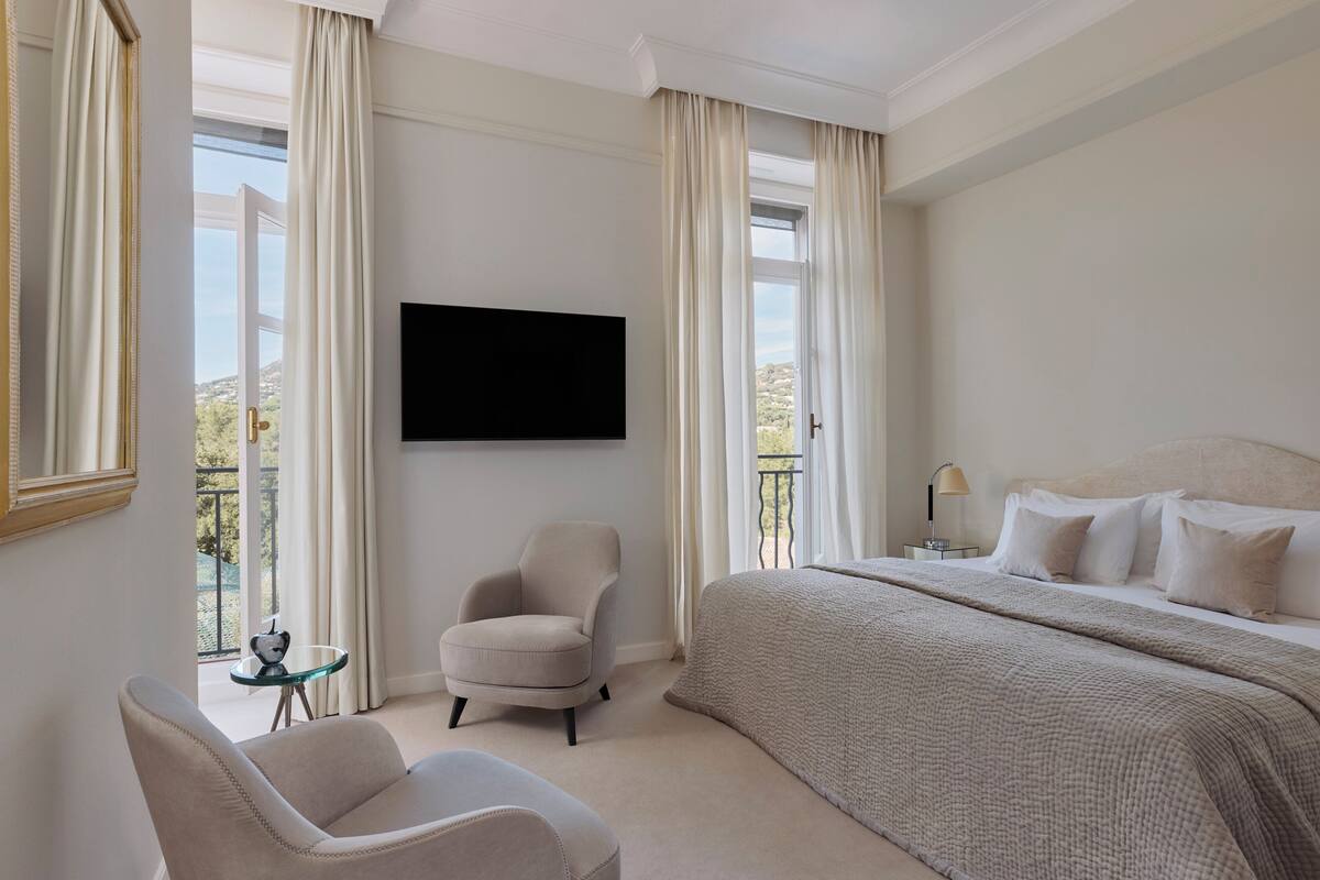 Une chambre d’hôtel avec un grand lit, un téléviseur fixé au mur, une chaise, une petite table et un balcon avec des rideaux.