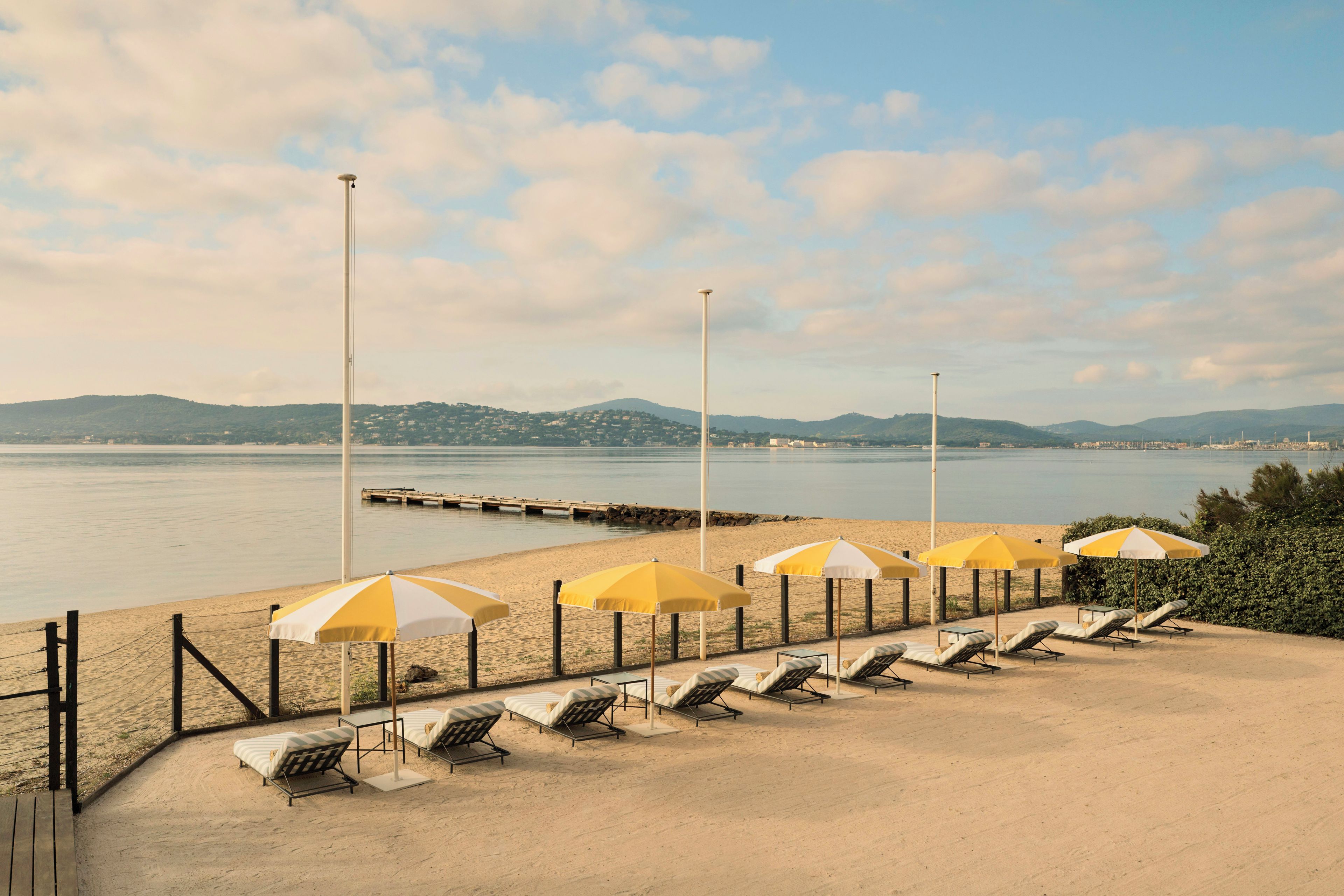 A beachside area with sun loungers and umbrellas, a wooden walkway, and a view of the sea and distant hills.