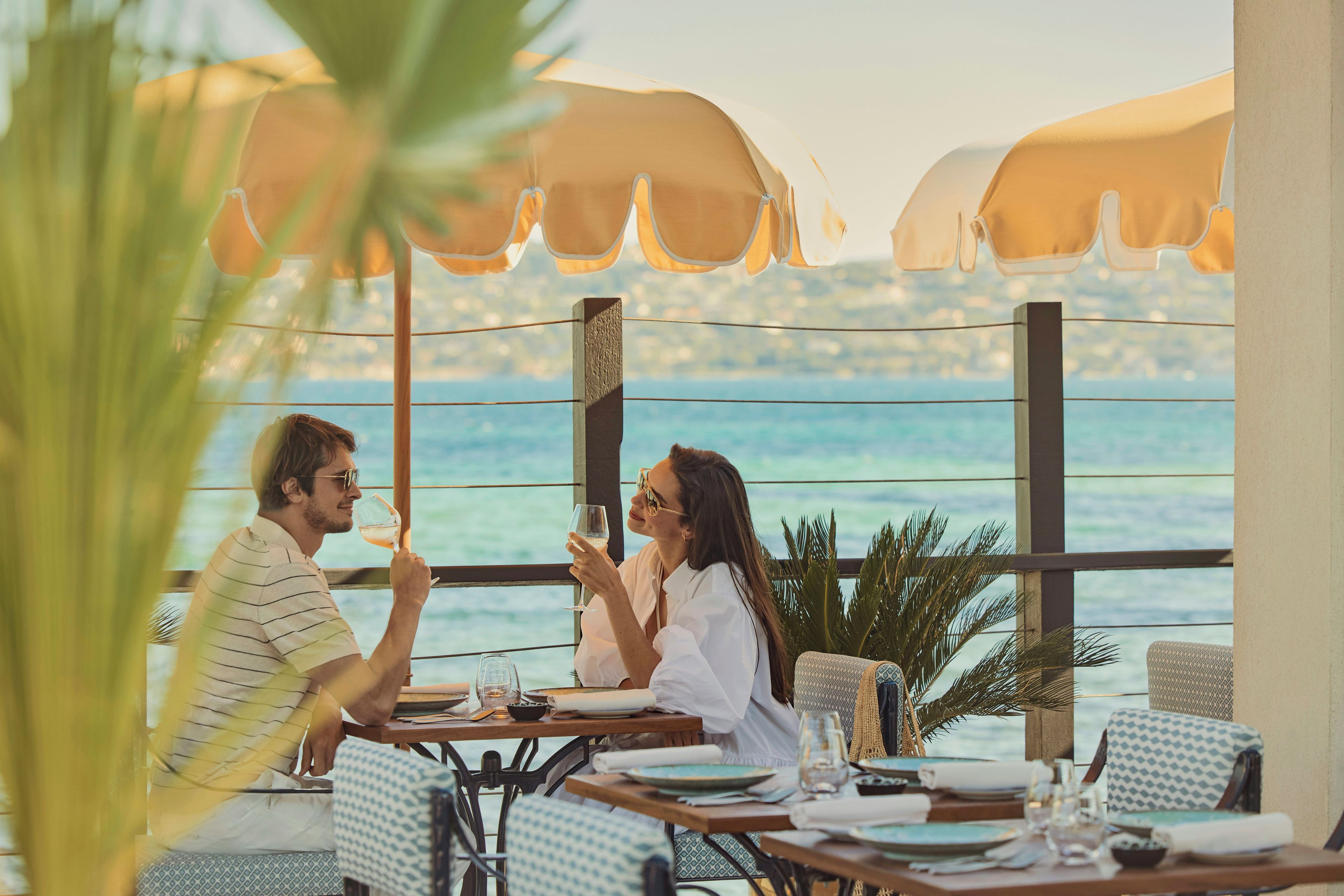 Un couple est assis à une table en plein air, avec vue sur la mer, protégé par de grands parasols. La table est dressée pour deux, avec des chaises.
