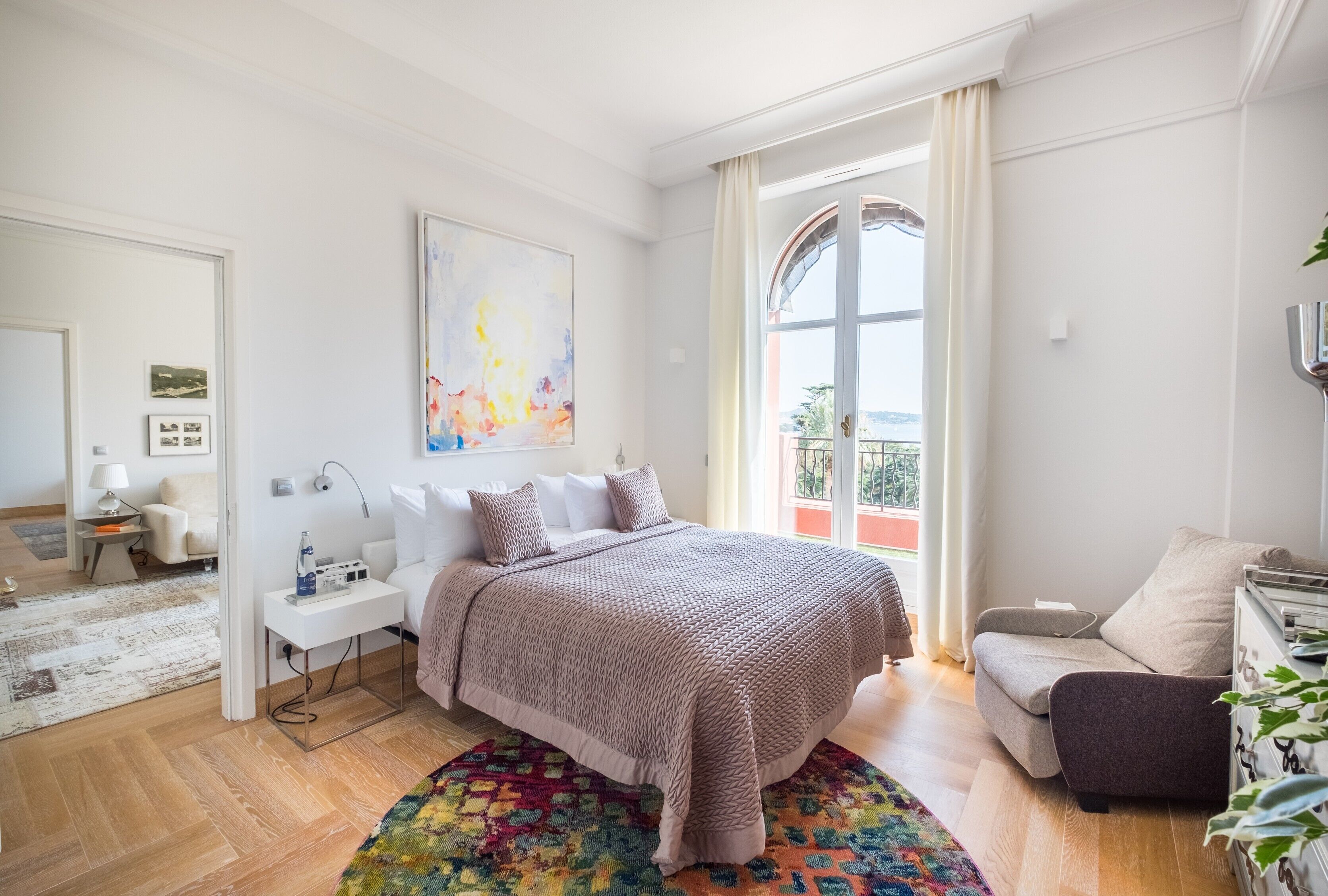 A modern bedroom with a large bed, a bedside table with a lamp, a colorful rug, and a view of the outdoors through a window.