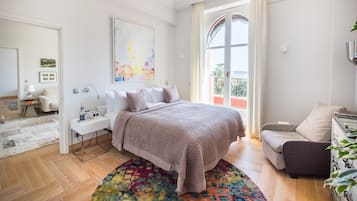 A modern bedroom with a large bed, a bedside table with a lamp, a colorful rug, and a view of the outdoors through a window.