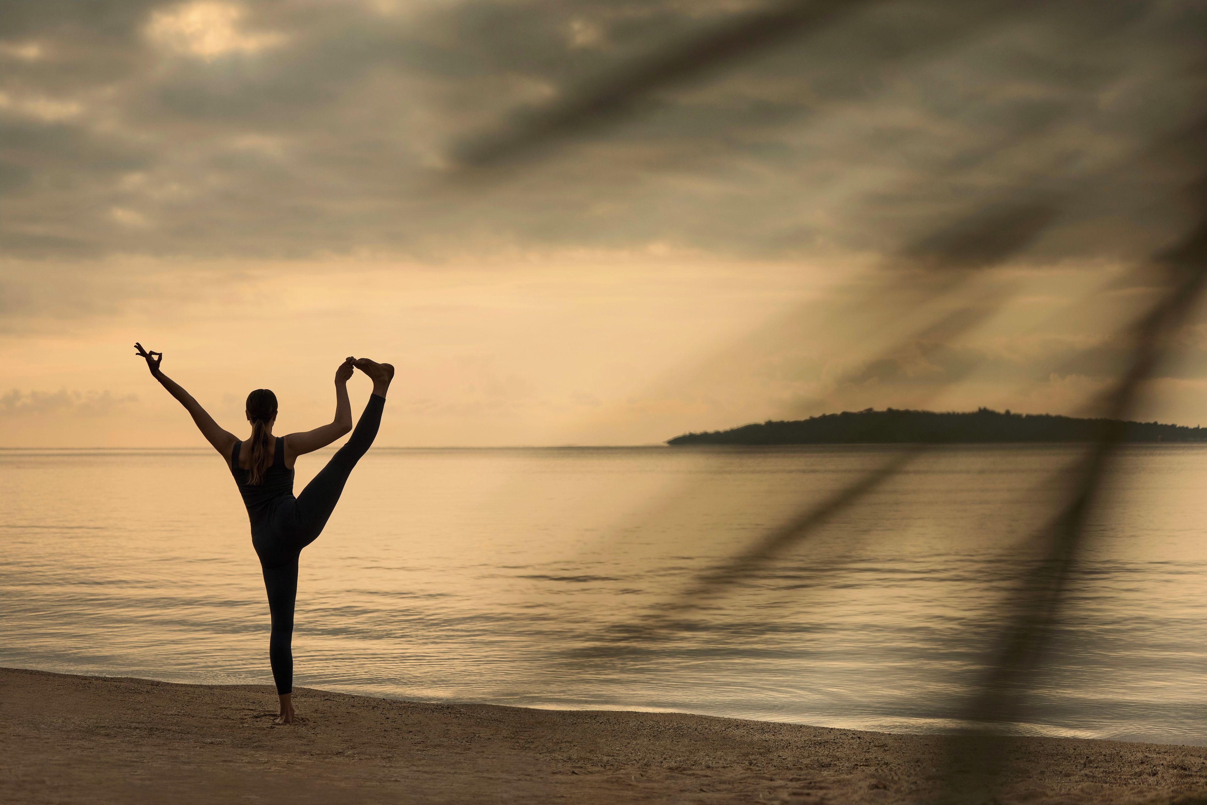 Una persona realizando una postura de yoga en la playa, con un mar tranquilo y un cielo nublado al fondo.