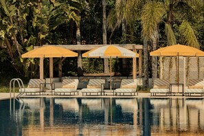 Zona junto a la piscina con sillones de descanso y sombrillas, rodeada de follaje tropical.