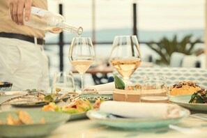 Une table à manger avec différents plats et verres, une personne en train de verser une boisson, et une vue sur la mer en arrière-plan.