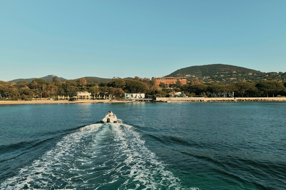 Ein Boot bewegt sich auf ruhigem Wasser, im Hintergrund eine hügelige Landschaft und Gebäude.
