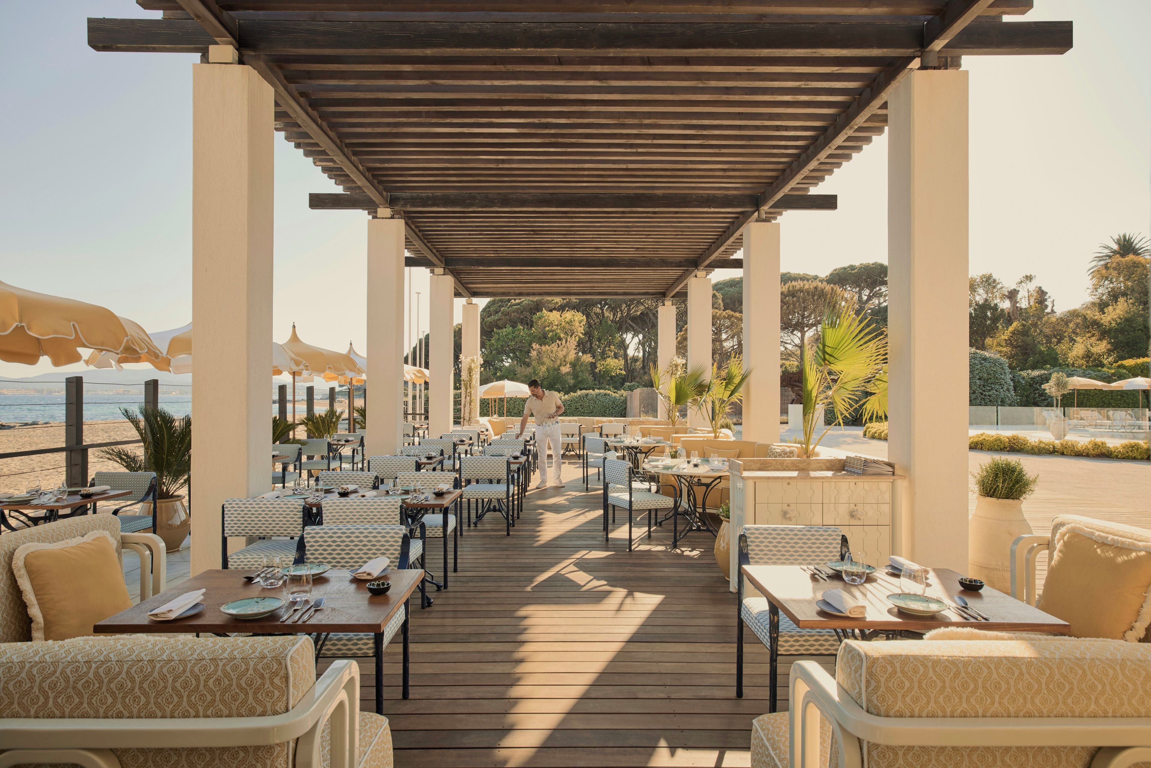 An outdoor dining area with tables and chairs, a wooden pergola, and a view of the beach.