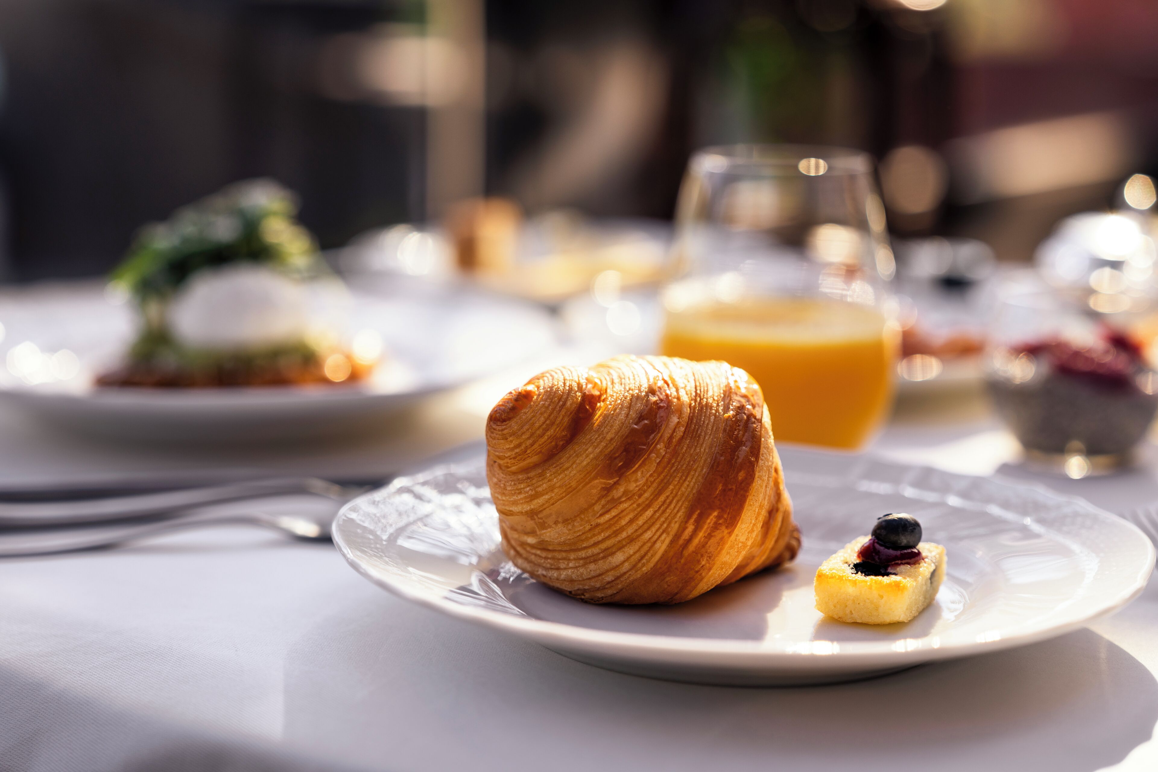 En frukostuppdukning med en croissant och en liten efterrätt på en tallrik, ett glas apelsinjuice och en skål med frukt i bakgrunden.