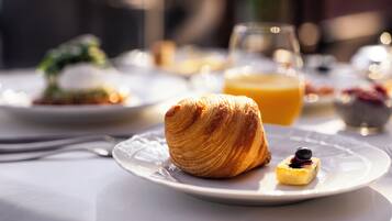 En frukostuppdukning med en croissant och en liten efterrätt på en tallrik, ett glas apelsinjuice och en skål med frukt i bakgrunden.