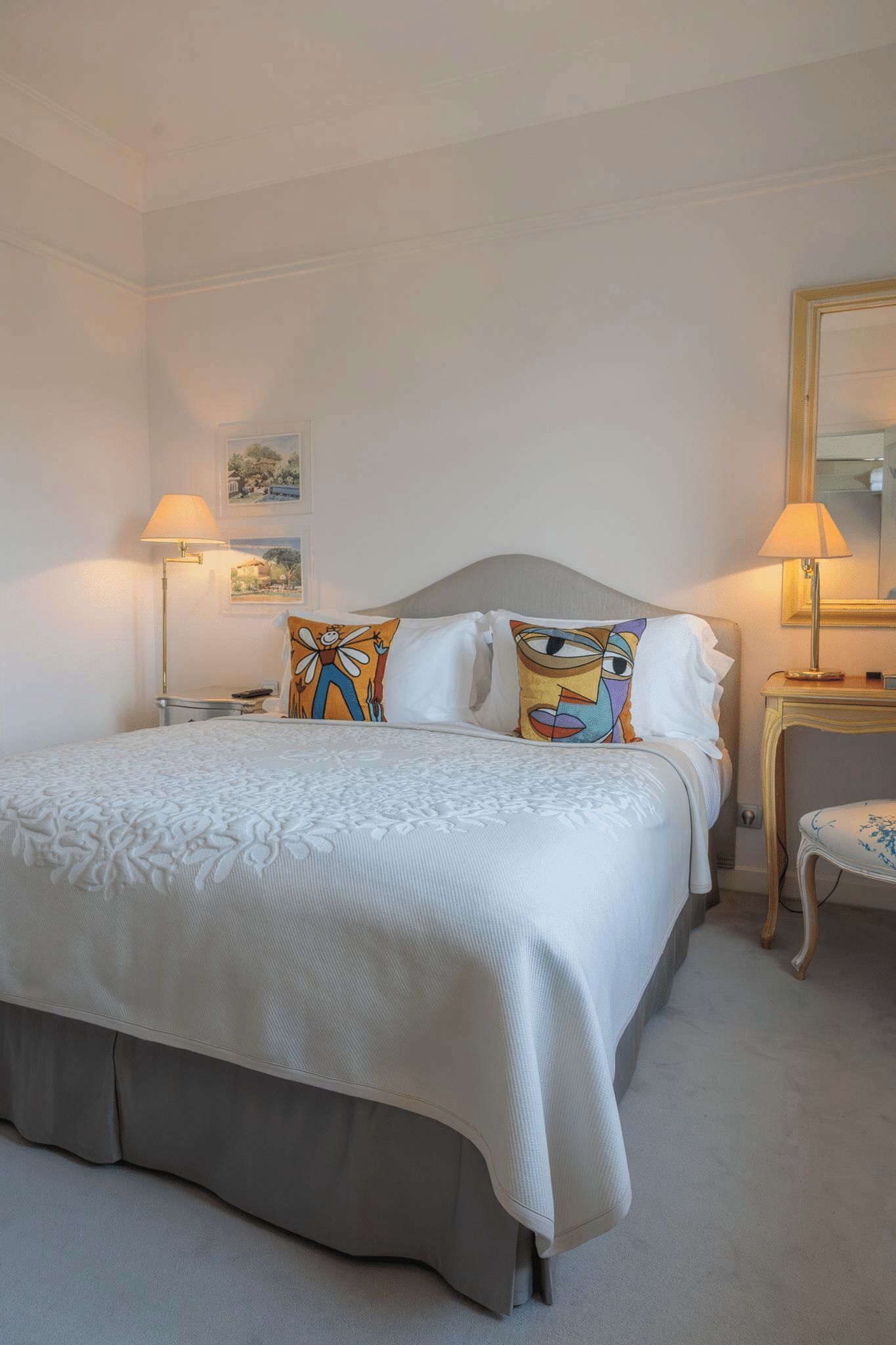 A neatly made bed with a grey headboard, white linens, and two decorative pillows. There are two bedside tables with lamps, a small chair, and a mirror on the right side.