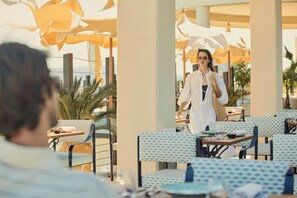 A person walking through a restaurant with outdoor seating, palm trees, and a view of the ocean. - COMO Le Beauvallon (Grimaud)