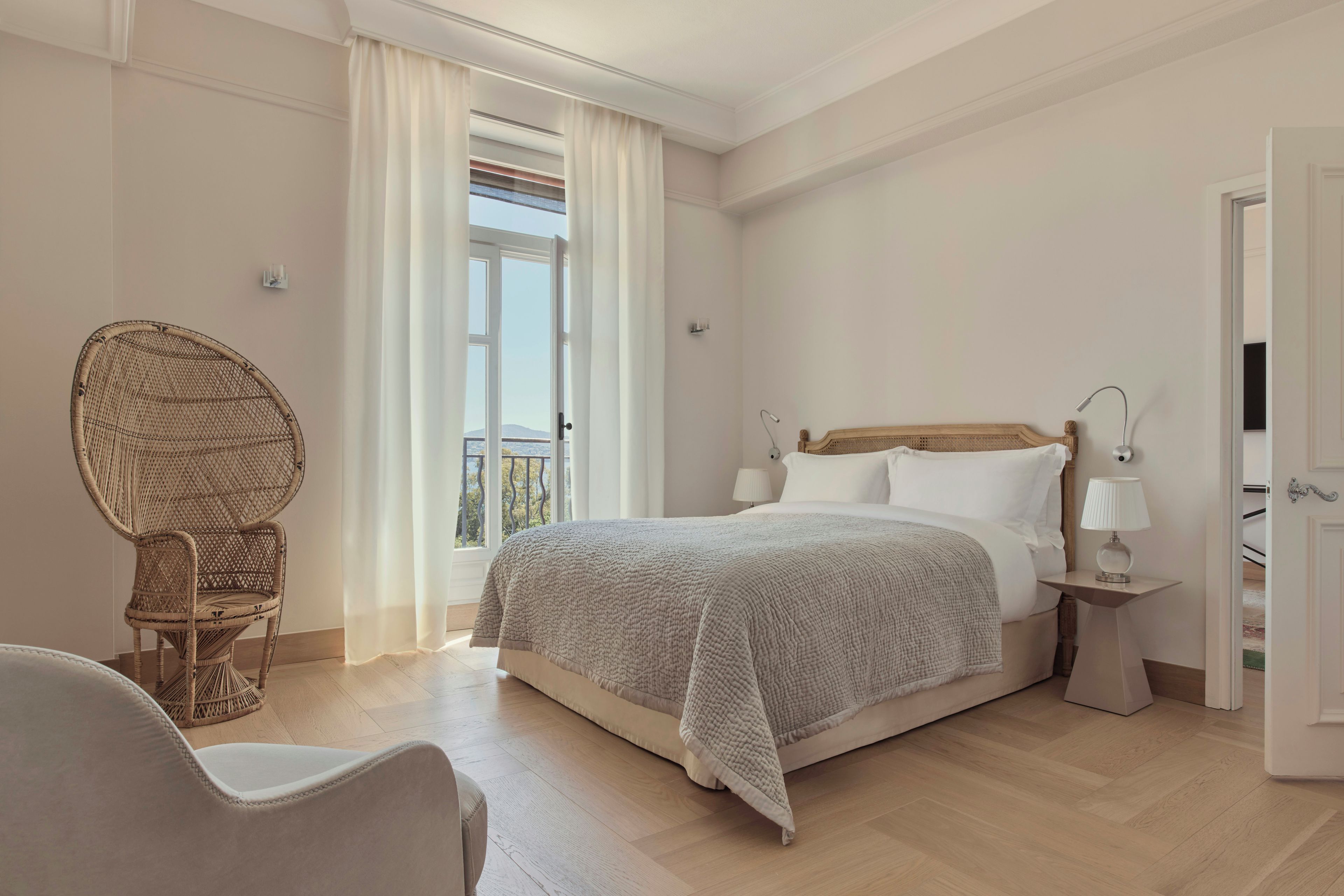 Un dormitorio con una cama grande, una silla de mimbre, una mesita de noche con lámpara y vistas al exterior a través de una ventana.