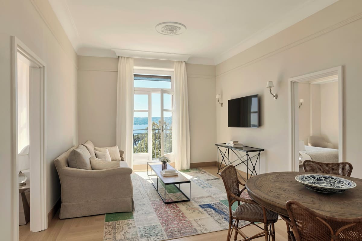 Un salon comprenant un canapé, une table basse, une table à manger avec des chaises, un téléviseur fixé au mur et une grande fenêtre avec des rideaux.