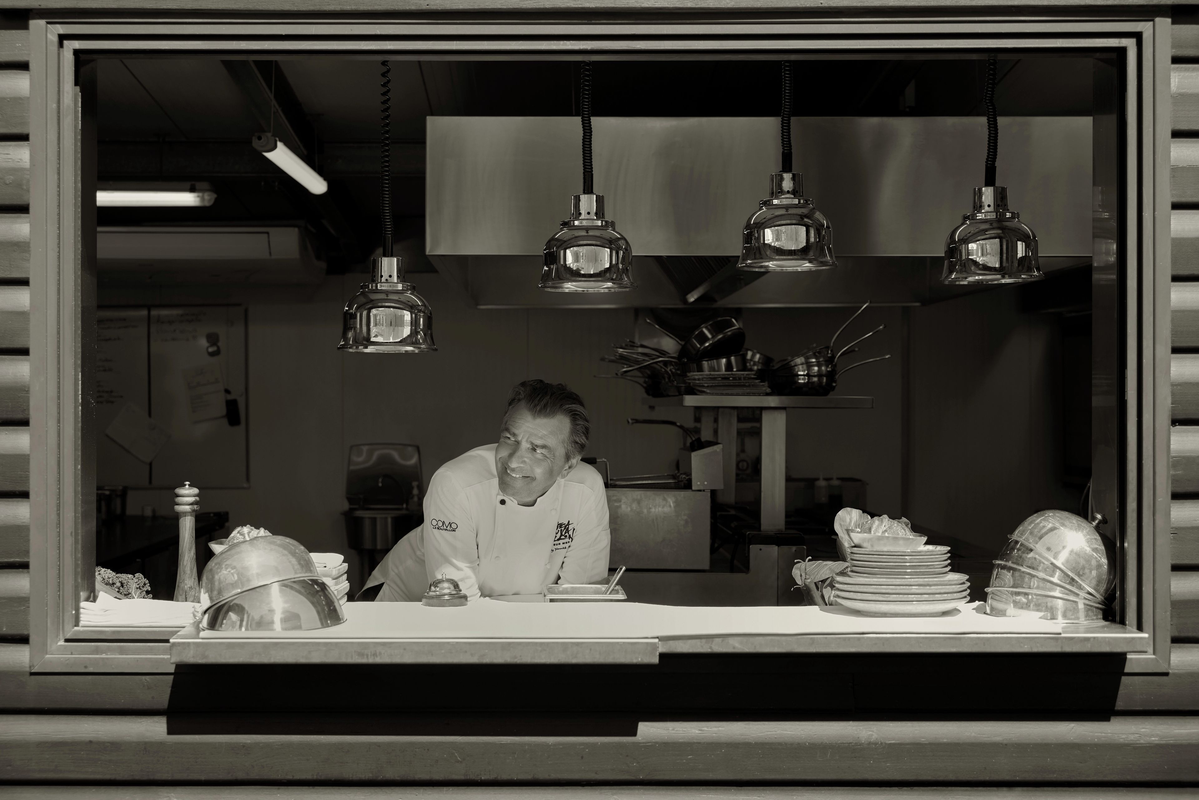 A chef at a counter with hanging lights, a stack of plates, and kitchen equipment in the background.