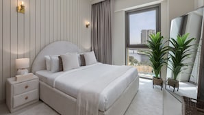 A modern bedroom with a large bed, bedside tables, a nightstand, a mirror, and two potted plants.
