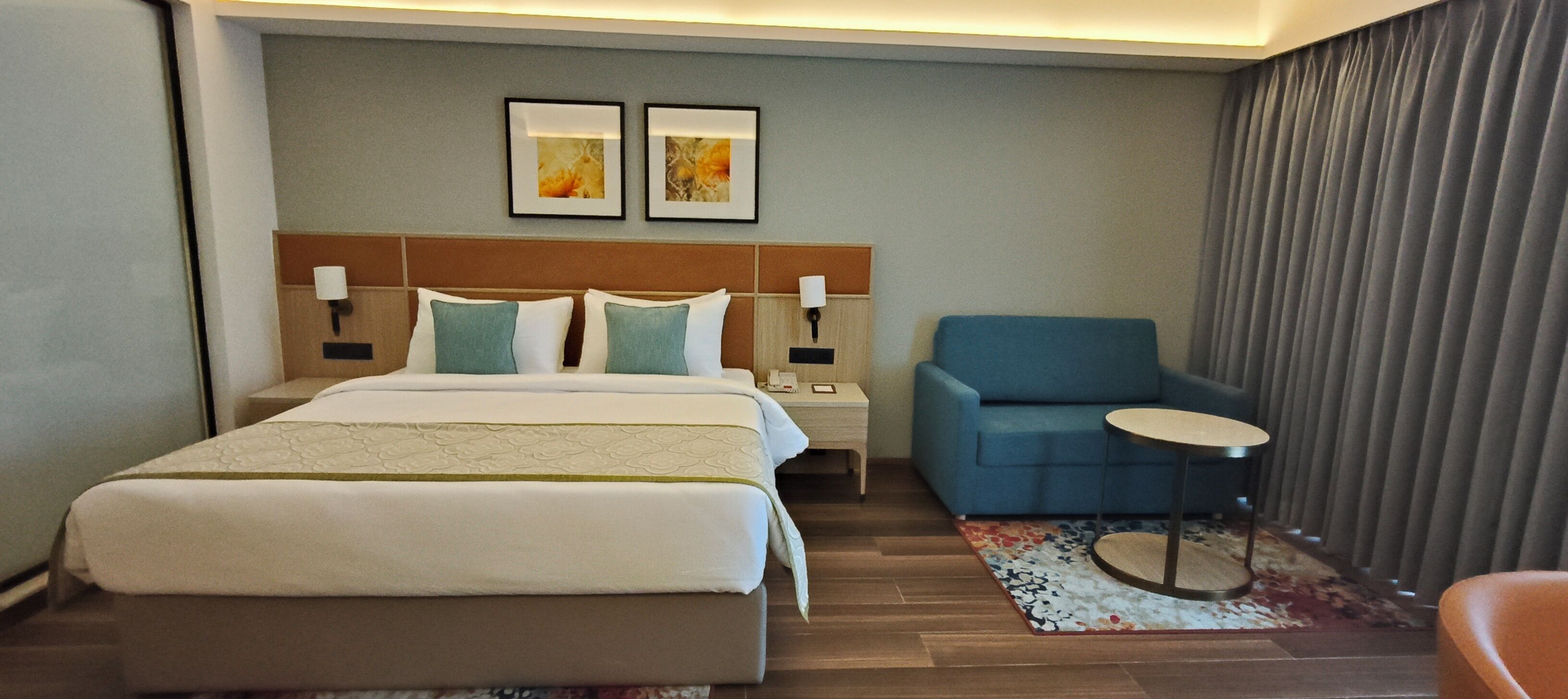 Una habitación de hotel con una cama grande, un sillón azul, una mesita redonda y dos cuadros en la pared.