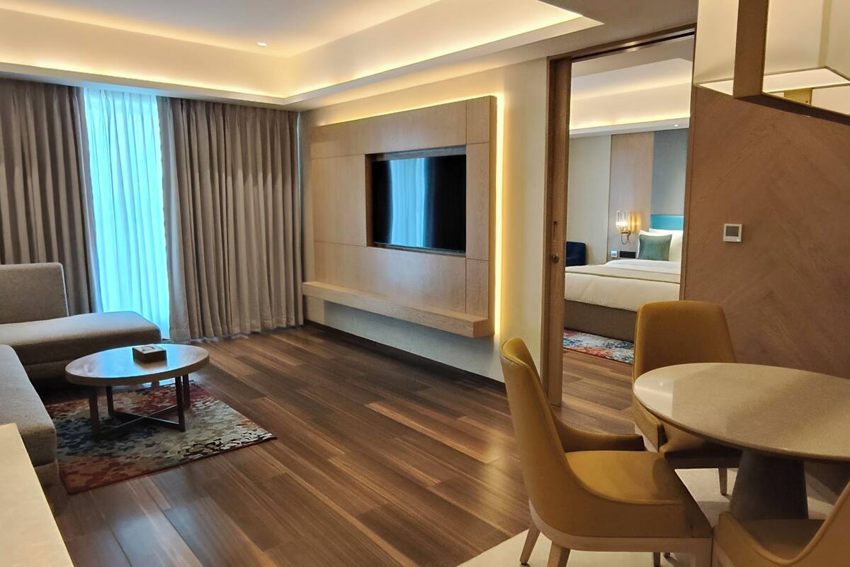 Ein modernes Hotelzimmer mit einer Couch, einem Esstisch mit Stühlen, einem an der Wand befestigten Fernseher und einem großen Fenster mit Vorhängen.