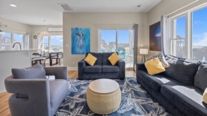 A modern living room with a grey sofa set, a round coffee table, and a large window with a city view. - Steps to Beach Access, 4 Bedrooms w/ King Suite, Fenced Yard, Pup Friendly (North Topsail Beach)