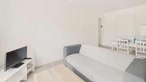 A modern, minimalistic bedroom with a grey sofa, a flat-screen TV on a white stand, and a white dresser with a mirror.