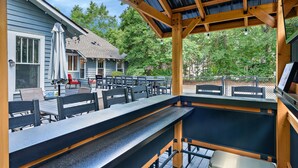 Eine Terrasse im Freien mit Holztischen und -stühlen, einem überdachten Sitzbereich und Blick auf ein Haus und Bäume.