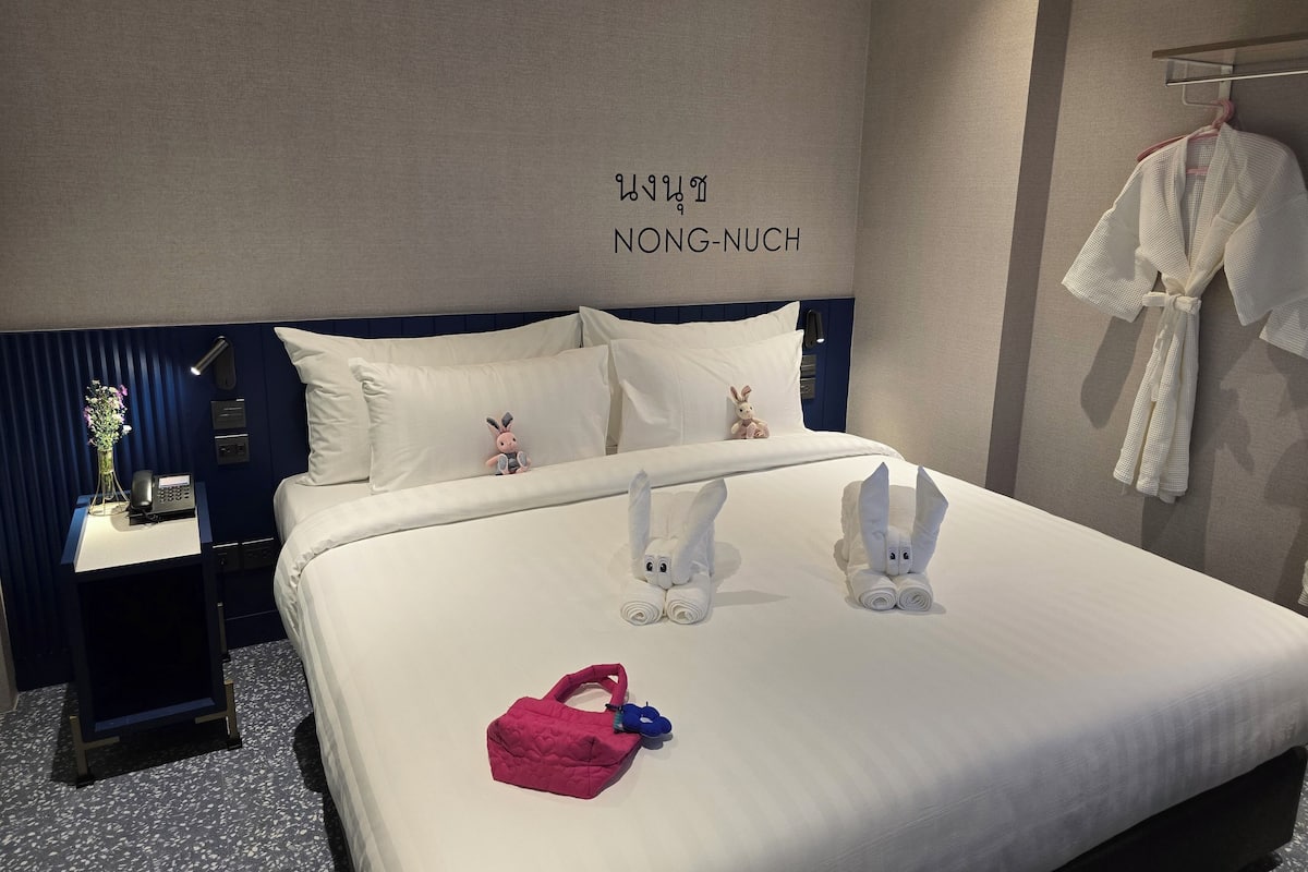 Un lit bien fait, avec une literie blanche, un sac rose et deux serviettes blanches en forme de lapins.