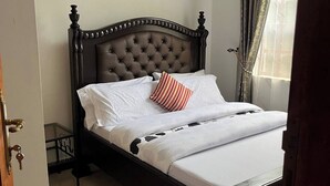 Una cama bien hecha con ropa blanca y una almohada a rayas, un cabecero de madera oscura y vistas a un edificio a través de una ventana con cortinas.