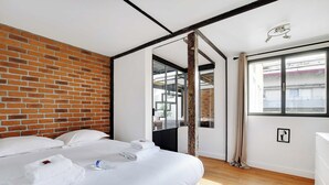 Une chambre d’hôtel avec un mur en briques, un grand lit avec du linge de lit blanc, un plancher en bois et une fenêtre avec des rideaux.