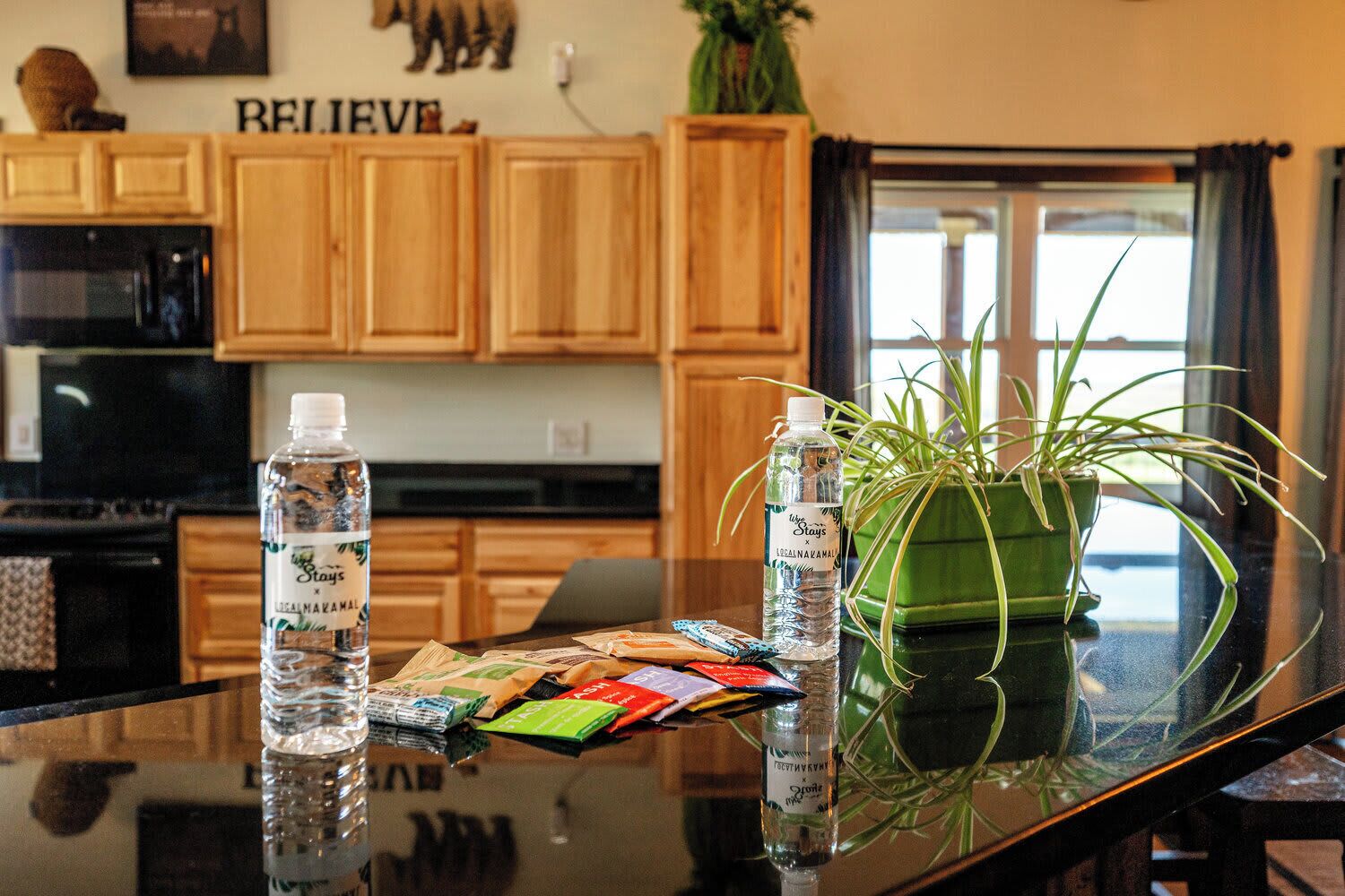 A kitchen with wooden cabinets, a microwave, a glass table with a plant and water bottles, and a laptop.