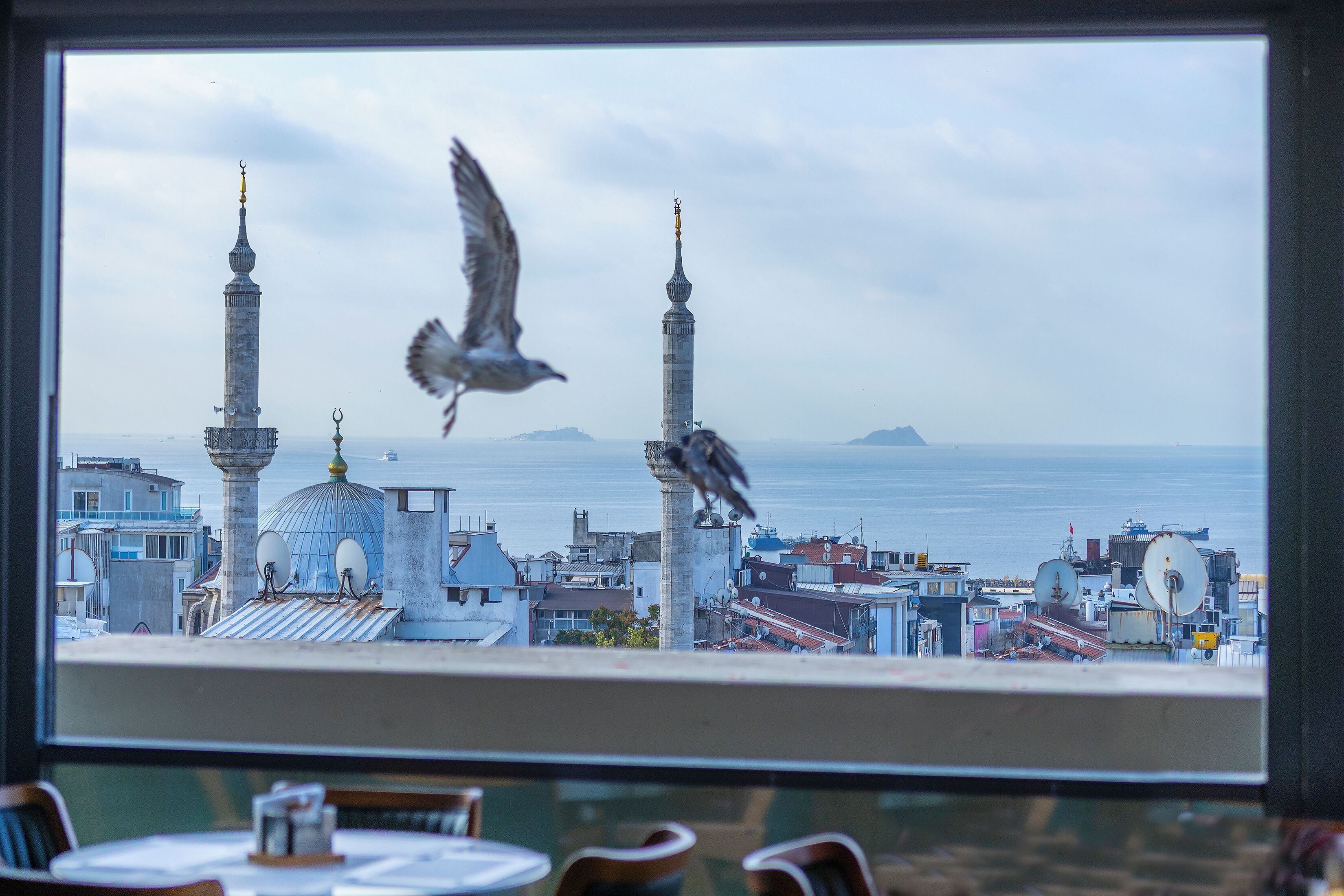 Une vue sur un paysage urbain avec une mosquée, dont on aperçoit les minarets et le dôme, à travers une fenêtre donnant sur une étendue d’eau.