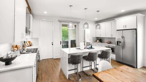 Una cocina moderna con armarios blancos, una isla central con taburetes de bar, electrodomésticos de acero inoxidable e iluminación colgante.