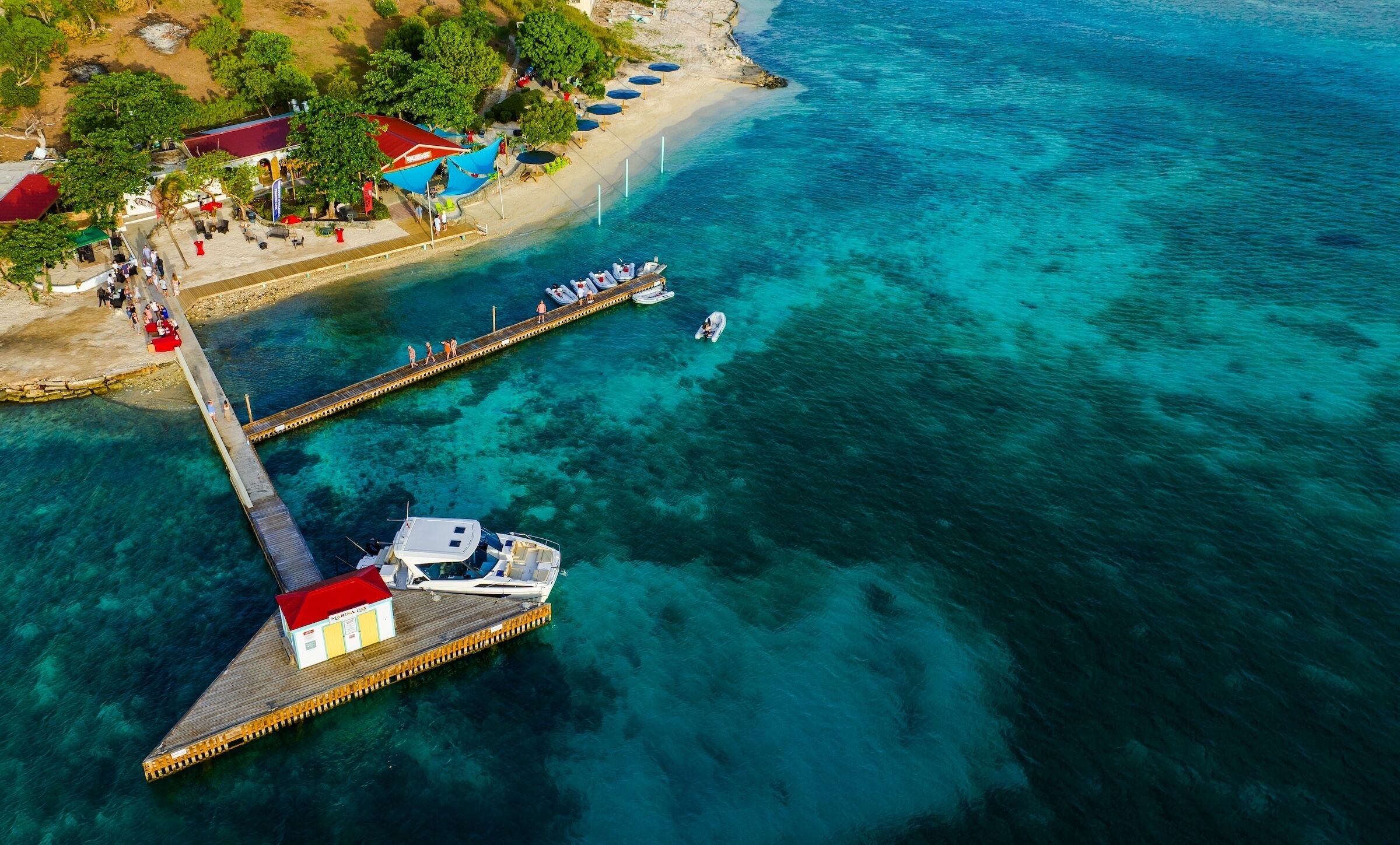 An aerial view of a coastal area with a pier extending into turquoise waters, a boat docked at the pier, and a beach with umbrellas and people.