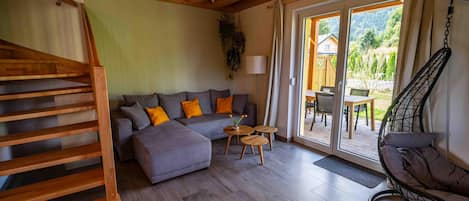 A living room with a wooden staircase, a grey sofa with orange cushions, a hanging chair, and a view of the outdoors.