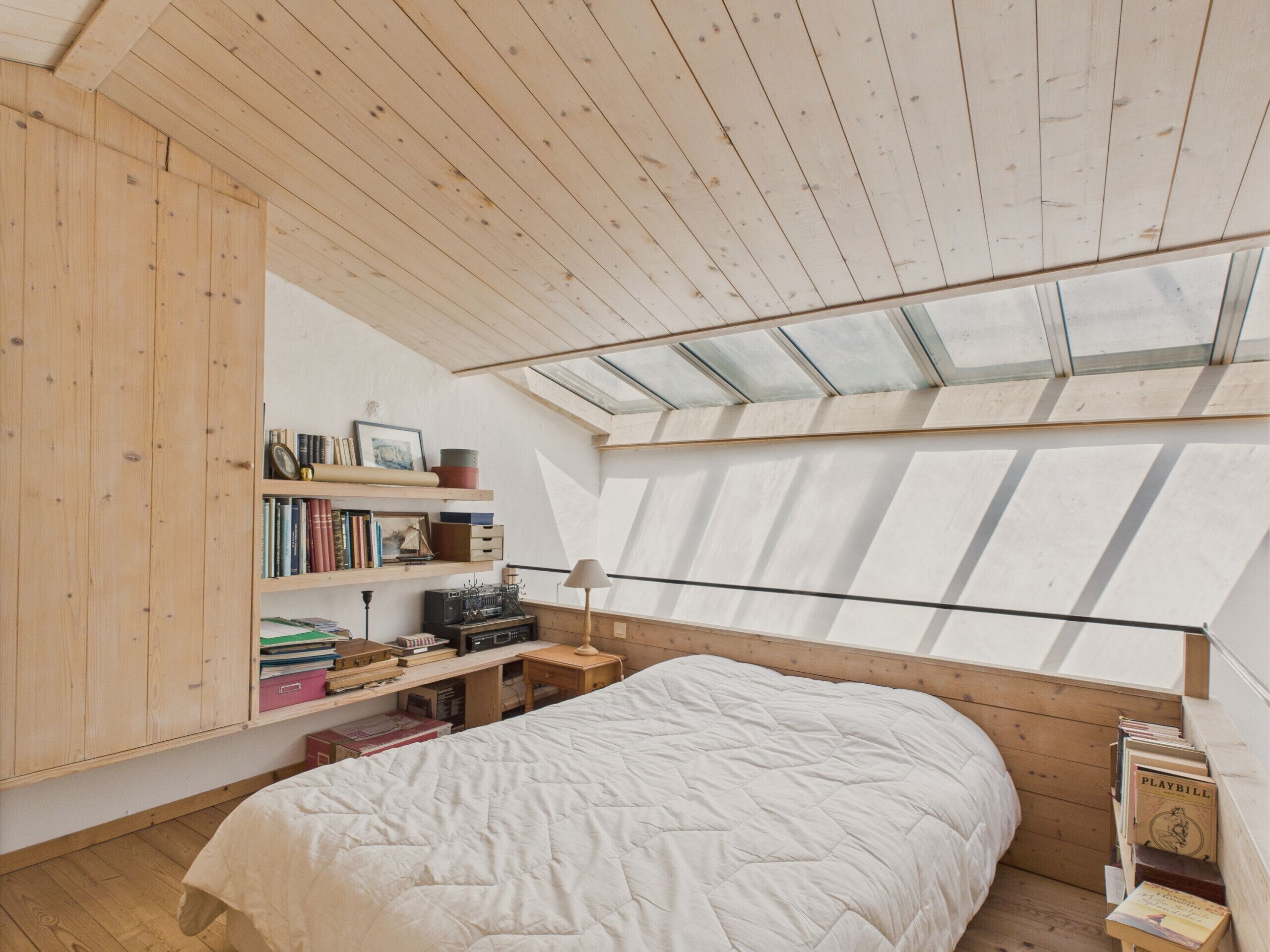 A bedroom with a sloped ceiling, skylight, wooden bookshelves, a bed, and a small table with a lamp.