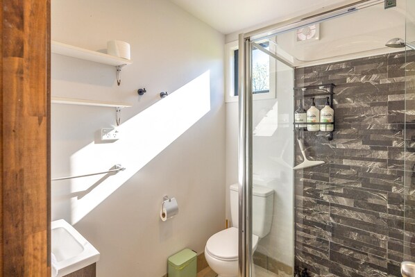 Un baño moderno con ducha de vidrio, inodoro, lavamanos y un estante con artículos de aseo.