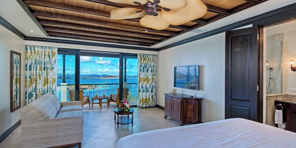 A hotel room with a large bed, a sofa, a small table with flowers, a ceiling fan, and a view of the sea through large windows.