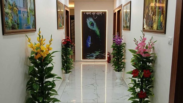 A hallway with white walls, marble flooring, and ceiling lights. There are potted plants and framed pictures on the walls. - Hotel RR Inn (Vadodara)