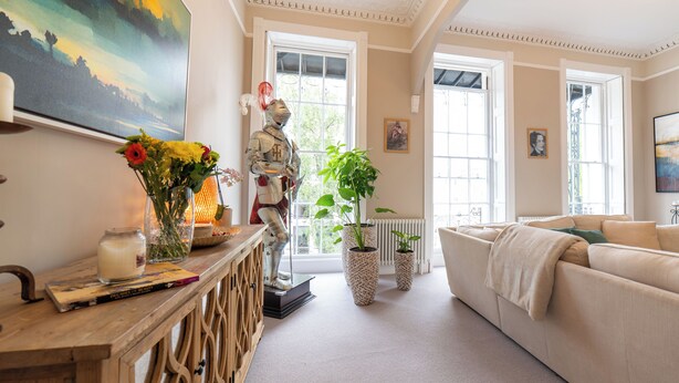 Ein helles Wohnzimmer mit einer Couch, einem Holztischchen, einer Vase mit Blumen und einer Statue. Große Fenster sorgen für natürliches Licht und einen Blick ins Freie.