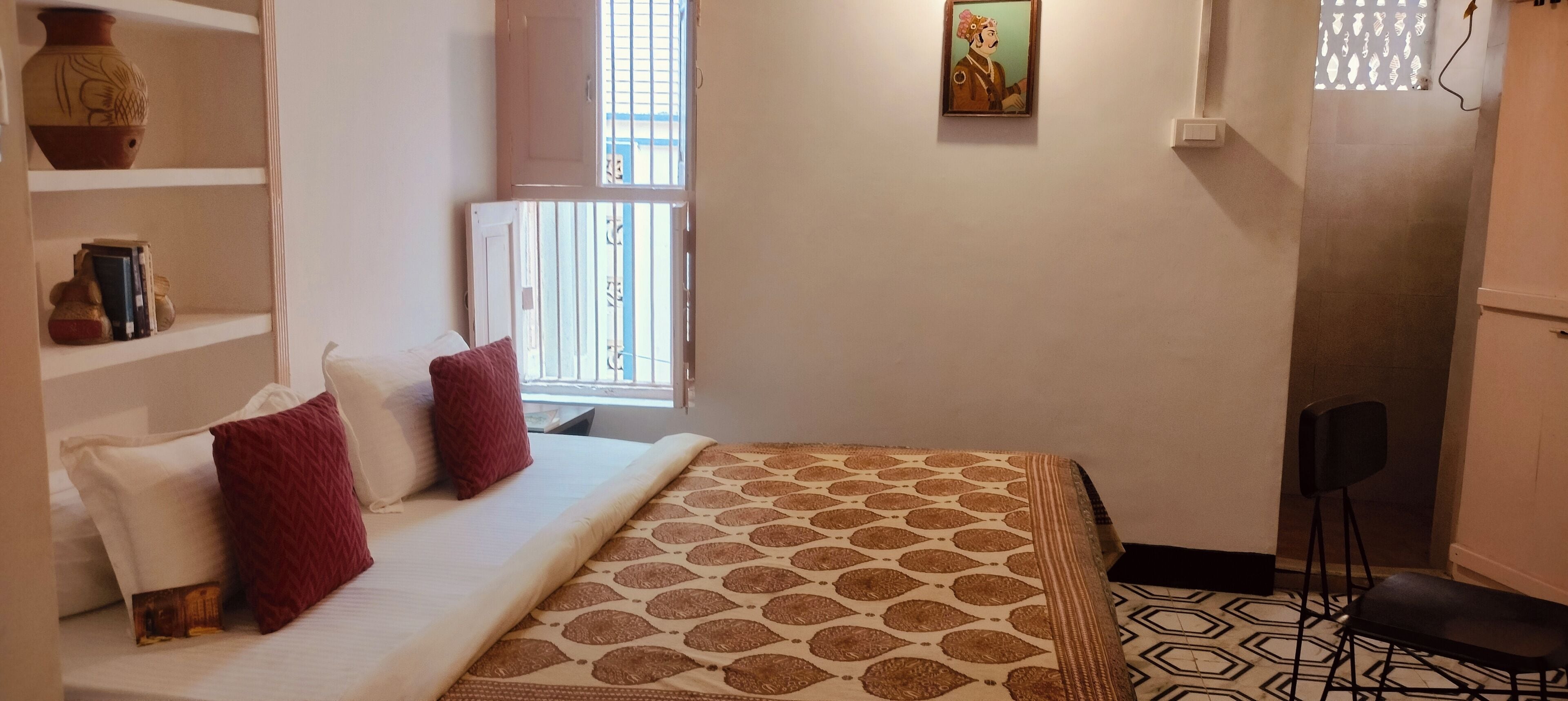A hotel room with a bed, a chair, a shelf with decorative items, and a window with a view.