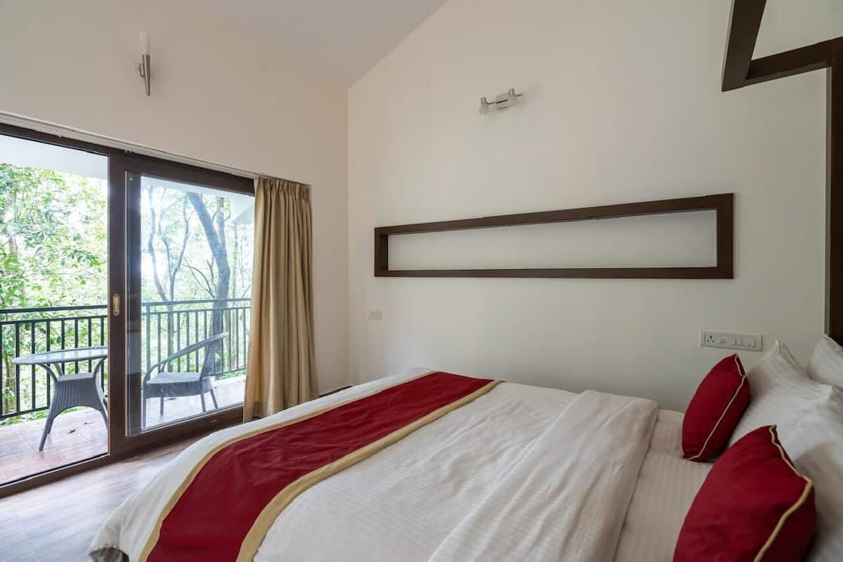 A hotel room with a bed, a window with curtains, a balcony with chairs, and a wall-mounted light fixture.