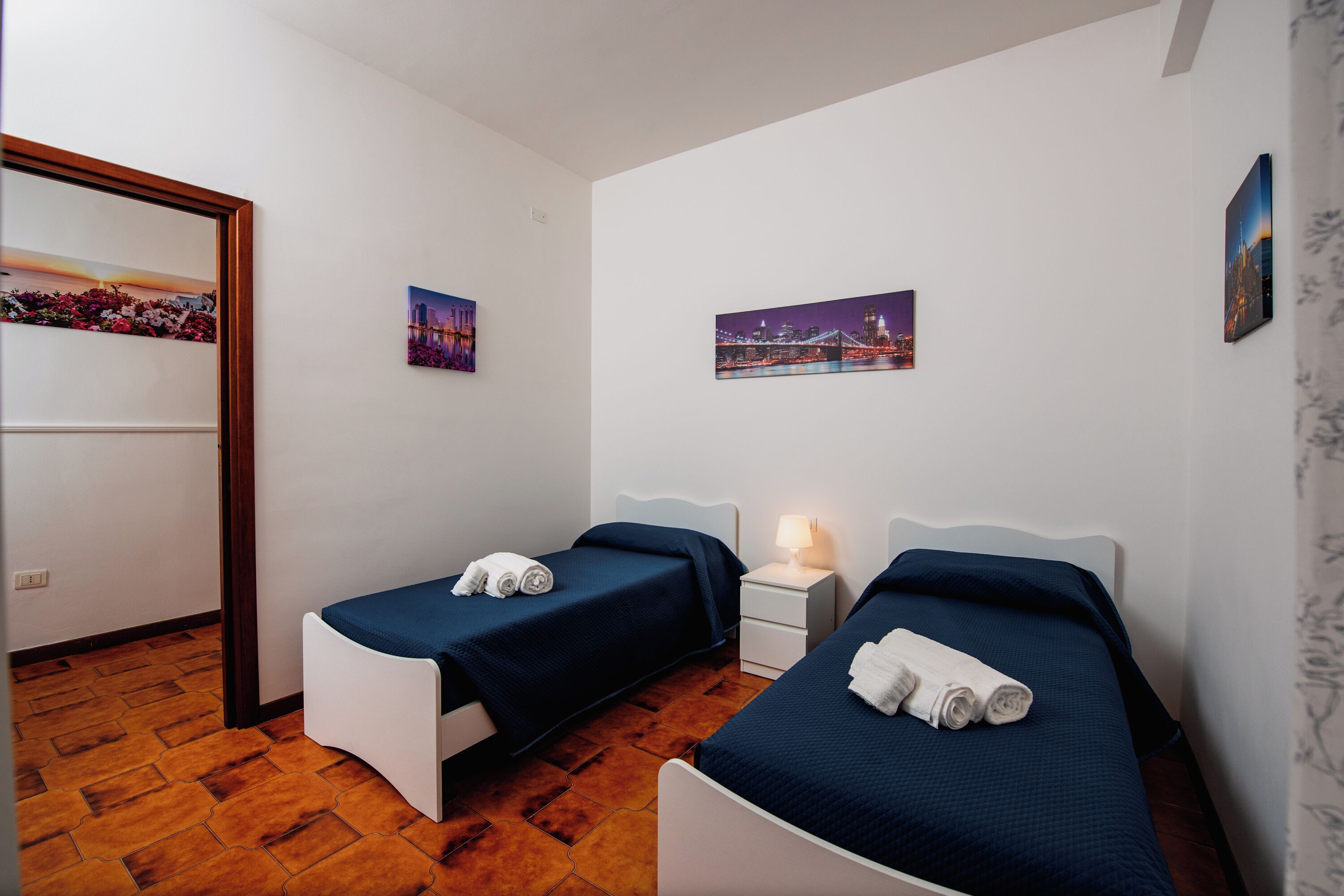 A hotel room with two single beds, a nightstand with a lamp, and two wall-mounted pictures.