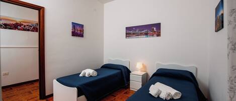 A hotel room with two single beds, a nightstand with a lamp, and two wall-mounted pictures.