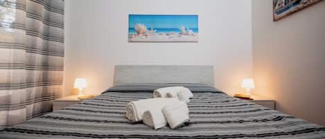 A neatly made bed with striped bedding, two bedside lamps, and a painting of a beach scene on the wall.