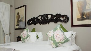 Una cama bien hecha con sábanas blancas y almohadas estampadas con motivos florales, un cabecero negro ornamentado y un cuadro colgado sobre la cama.